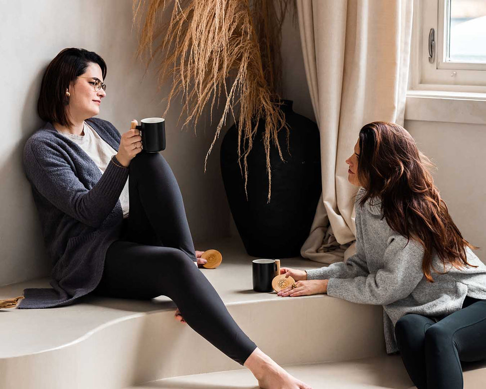 Two young women with dark hair in a bright living space sit and talk while drinking tea